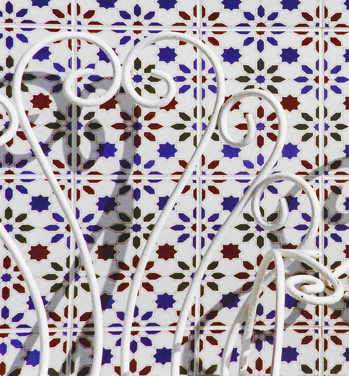 Detalle macro de forja blanca en espiral con sombra proyectada sobre azulejos mediterráneos en azul cobalto, burdeos y verde, patrón geométrico levantino IMAJIN-ARTE
